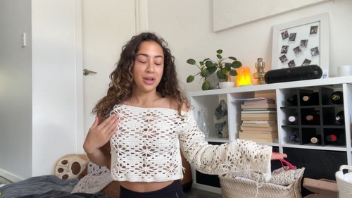Tutorial presenter holding up a finished cream crochet daisy flower top with long sleeves in a bright room, showing the front of the motif-join fabric
