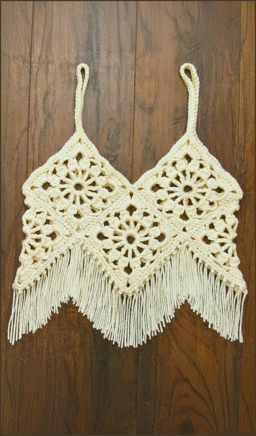 Flat lay of a cream crochet boho crop top made from joined open floral medallion squares with long fringe along the zigzag hem and loop spaghetti straps