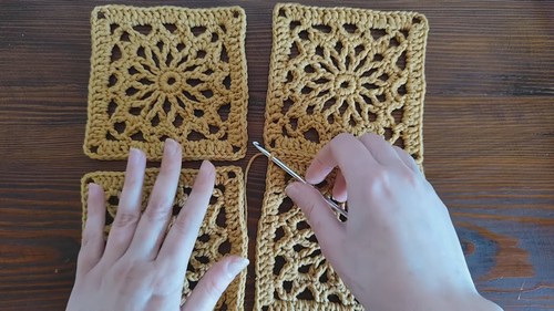 Hands joining four golden floral crochet squares together with a silver hook, showing the intricate lace medallion centers