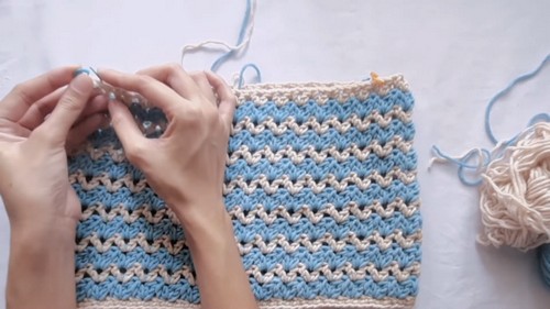 Close-up of hands crocheting into a blue and cream striped crochet fabric panel with a silver hook