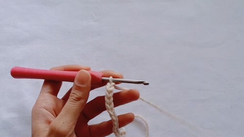 Hand holding a pink crochet hook with a short foundation chain of cream yarn against a white background 
