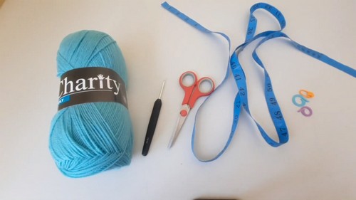 Supply flat lay showing a skein of teal Charity yarn, a small black crochet hook, red scissors, blue measuring tape, and two stitch markers 