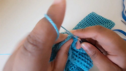 Close-up of hands working teal yarn with a small silver crochet hook into consistent rows of stitches 