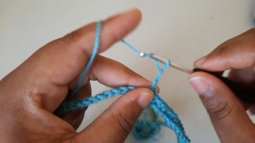Hands holding a foundation chain of bright teal yarn with a small silver crochet hook 