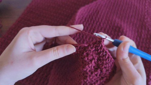 Close-up of hands working a blue crochet hook into a dark burgundy star stitch fabric, showing the raised bumpy stitch texture 