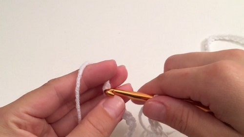 Hands forming early chain stitches with white yarn and a gold crochet hook against a white background. 