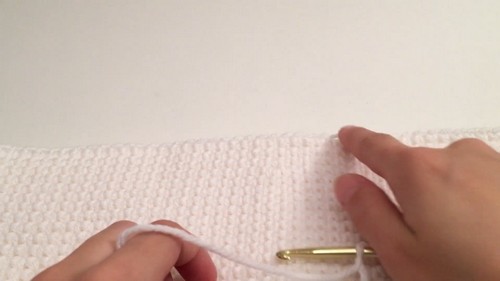 Hands working a gold crochet hook along the edge of the white dense fabric, showing finishing or seaming work on the tank top. 