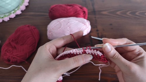 Hands working the first rows of a two-color vertical brick crochet pattern with a long silver hook, crimson and pink yarn balls visible in the background