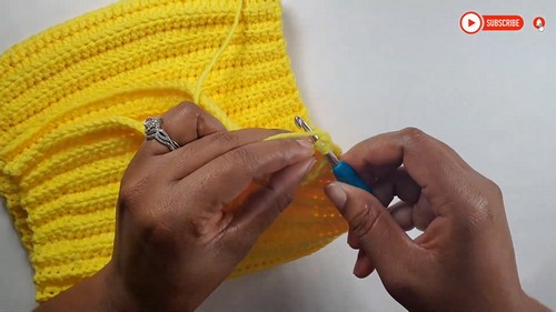 Hands working a blue crochet hook into the edge of a yellow ribbed crochet panel, showing a stitch step in the tube top construction with the rib columns visible in the background 