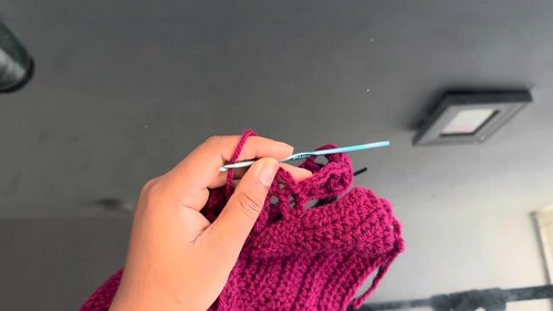 Hand holding a light blue crochet hook working into a section of burgundy ribbed crochet fabric 