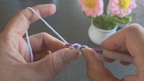 Hands forming the first chain stitches of a white crochet project with a purple hook, small fabric flowers visible in the background.