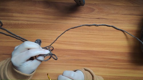 Hands forming a magic ring with dark gray yarn and a gold hook on a wooden surface