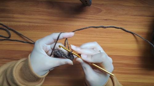 Hands crocheting the beginning of a brown yarn panel with a gold hook on a wooden surface