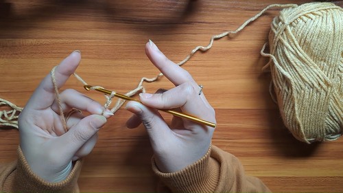Hands working a gold hook into a cream foundation chain on a wooden surface with a yarn skein nearby