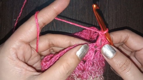 Close-up of hands using an orange crochet hook to work a stitch into a pink variegated crochet fabric, slightly different angle