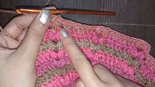 Hands working a pink and brown variegated crochet swatch showing shell or cluster stitch rows, orange hook inserted at top