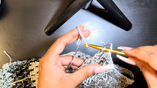 Working the grey and white variegated section of the cold shoulder sweater with a gold hook on a dark desk surface.