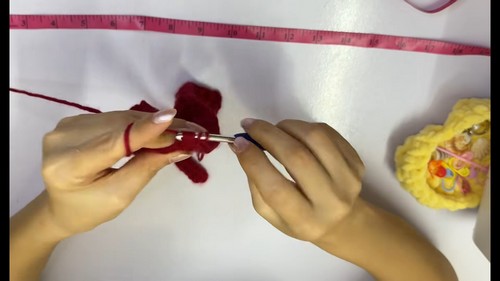 Hands working the foundation chain of a red crochet project with a blue hook, red yarn and measuring tape visible on a white surface