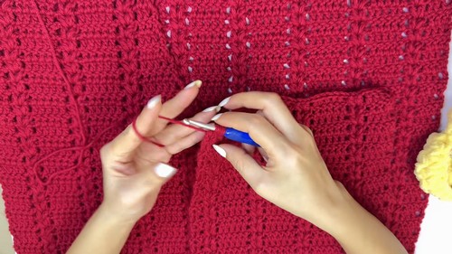 Hands using a blue hook to crochet along the body of a completed red sweater laid flat, showing the repeating stitch texture across the fabric