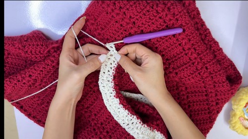 Hands working a white lace collar border onto a red crochet sweater with a purple hook, open shell lace edging curving along the neckline
