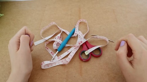 Hands resting beside a teal crochet hook, red scissors, and a coiled measuring tape on a tan surface.