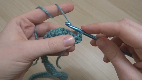 Hands forming the foundation of a teal crochet strip with a gray hook showing the first few stitches