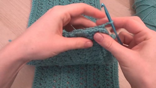 Hands using a blue hook to pick up stitches along the edge of a teal crochet panel for the cap sleeve border