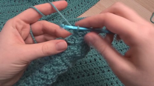 Hands working a blue hook through the textured rows of a teal crochet panel showing the lace stitch detail