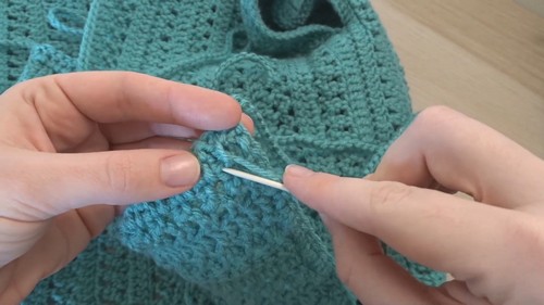 Hands using a tapestry needle to seam the shoulder edge of a teal crochet rectangle panel