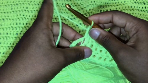 Hands working a neon green stitch with a gold hook along the open mesh body of a crochet sweater.