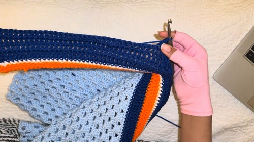 gloved hand holding up a partially assembled navy and light blue sweater panel showing the granny stitch body section and orange stripe