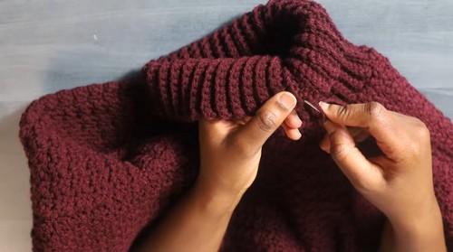 Flat lay of a burgundy crochet funnel neck sweater with hands weaving the yarn tail into the ribbed cuff.