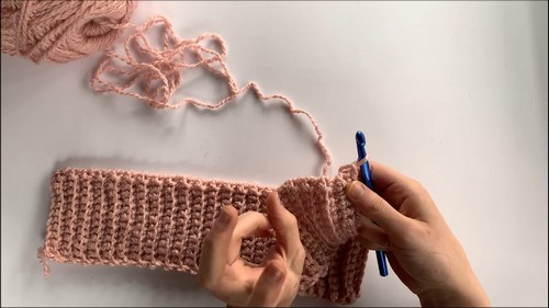 Hands working a ribbed crochet strip with a blue hook showing the foundation rows of the cuff or border section in pink yarn