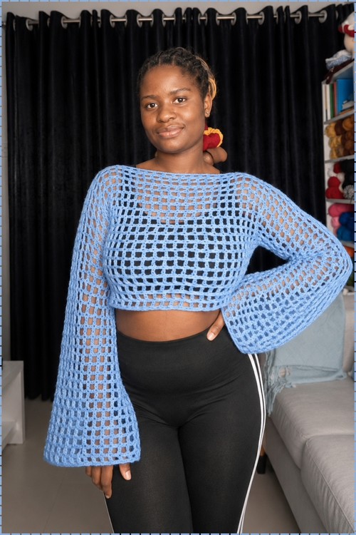 Finished crochet bell sleeve sweater in blue worn in a home studio, showing the wide boat neckline and full length of the flared mesh sleeves.