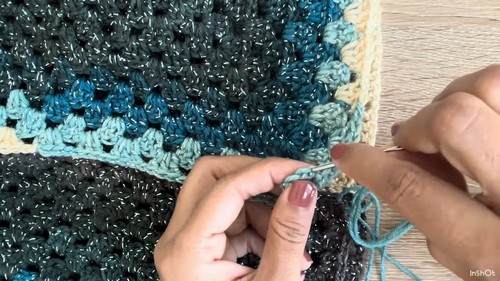 Attaching the teal accent edging at the neckline of the dark grey crochet sweater body, sleeve panel visible alongside.