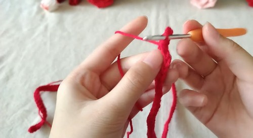 Hands working a foundation chain in red yarn with an orange crochet hook, showing the very start of the sweater pattern construction.
