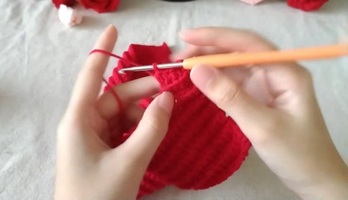 Hands using an orange crochet hook to work into the edge of a red crocheted ribbed panel, showing the vertical rib texture developing in the early body section.