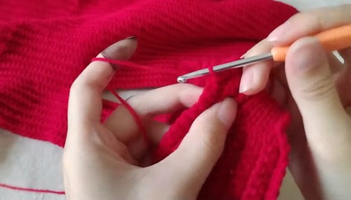 Hands working an orange crochet hook through a section of the red ribbed sweater fabric, showing a joining or construction step partway through the garment build.