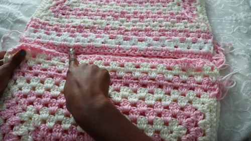 Hands joining two pink and white granny stitch sweater panels together with a crochet hook, work laid flat on a bed