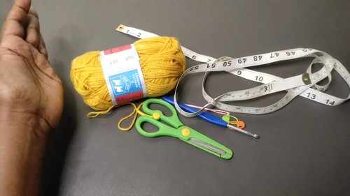 Yellow yarn skein with tape measure scissors and crochet hooks laid out on a gray surface
