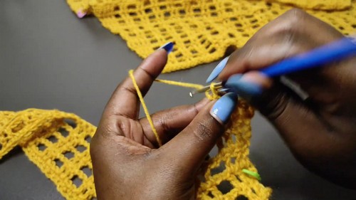 Hands using a blue hook to work into a yellow crochet mesh panel with a partially finished vest section visible behind