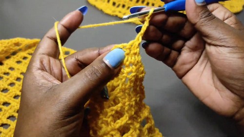 Hands working a blue hook through yellow yarn building the v stitch mesh body of the vest
