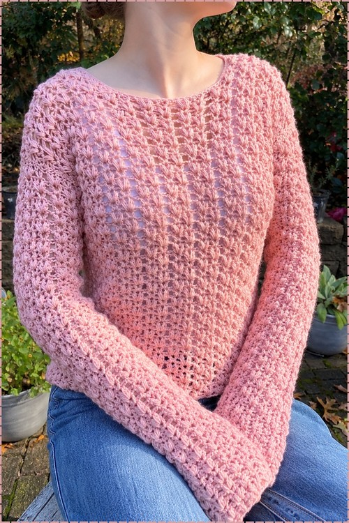 Woman wearing a pink long sleeve crochet sweater with an open puff V stitch texture seated outdoors in a garden setting
