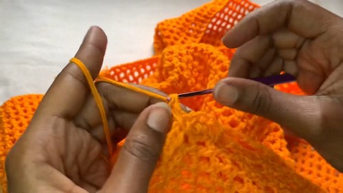 Working the open mesh body of the orange bell sleeve sweater with a purple hook, the grid structure of chain spaces clearly visible.