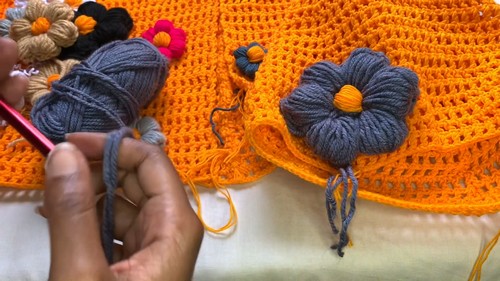 Making a grey puff flower appliqué in progress with finished flowers and the orange sweater panel visible in the background.