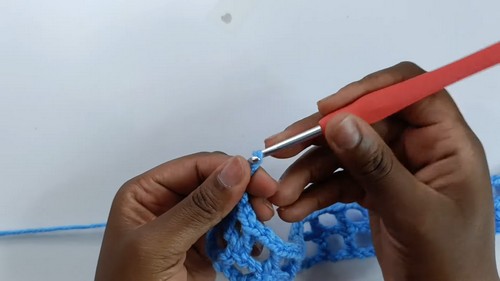 Working the early rows of the blue mesh sweater with a salmon pink hook, open grid structure visible in the growing fabric.
