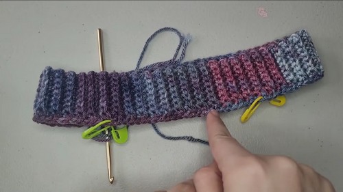 A ribbed variegated crochet neckband with yellow and green stitch markers clipped along the edge and a gold hook resting on top.