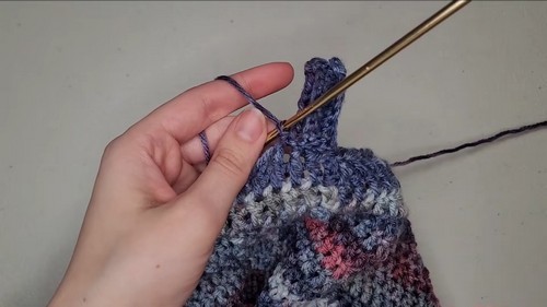 Hand guiding a gold crochet hook through the raglan increase point of a blue and pink variegated sweater in progress.