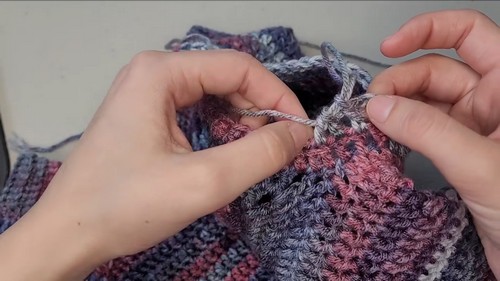 Hands joining the raglan seam of a blue and pink variegated crochet sweater with fingers guiding the stitch.