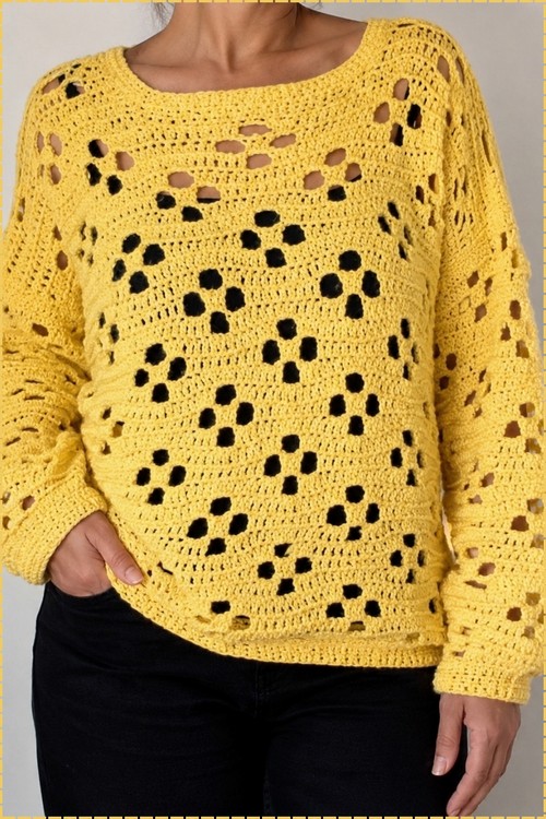 Woman wearing a yellow crochet scoop neck sweater with a floral eyelet body over black pants against a gray background.